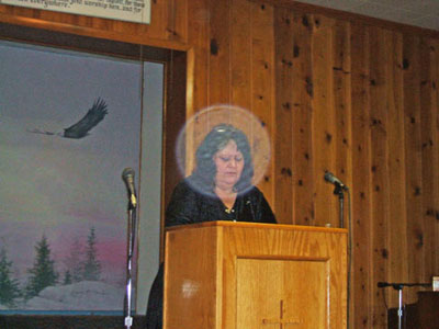 Woman speaking at a lectern in a wood-paneled room, with a circular spot over her face