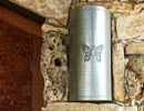 Cylindrical metal fixture with a butterfly emblem mounted on a stone wall