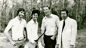 Four men standing together outdoors in an archery-club photograph